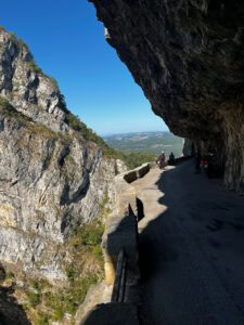 Eine Bergstraße schlängelt sich an einer felsigen Klippe entlang, während Menschen zu Fuß oder mit dem Fahrrad unterwegs sind; links ein steiler Abgrund, darüber der Himmel und die Aussicht in die Ferne.
