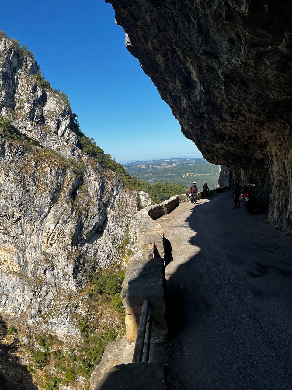 IMG_7682 Eine Bergstraße schlängelt sich an einer felsigen Klippe entlang, während Menschen zu Fuß oder mit dem Fahrrad unterwegs sind; links ein steiler Abgrund, darüber der Himmel und die Aussicht in die Ferne.