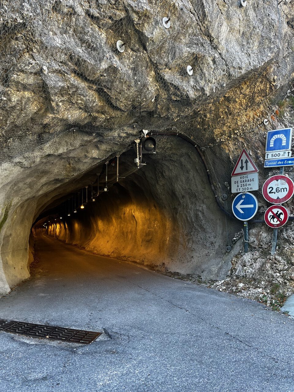 IMG_7685 Ein schmaler, beleuchteter Tunnel durch einen felsigen Hang mit mehreren Eingangsschildern, auf denen Höhen- und Breitenbeschränkungen sowie Pfeile angegeben sind.