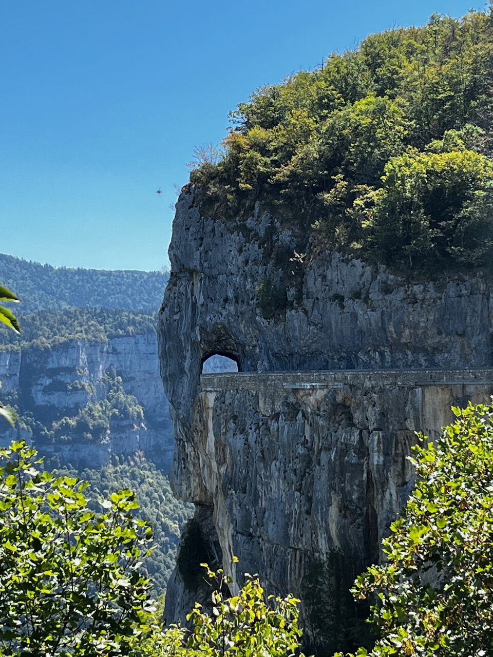 IMG_7691 Eine schmale Straße schlängelt sich durch einen Tunnel in einer felsigen Klippe, gesäumt von grünen Wäldern und Bergen unter blauem Himmel.