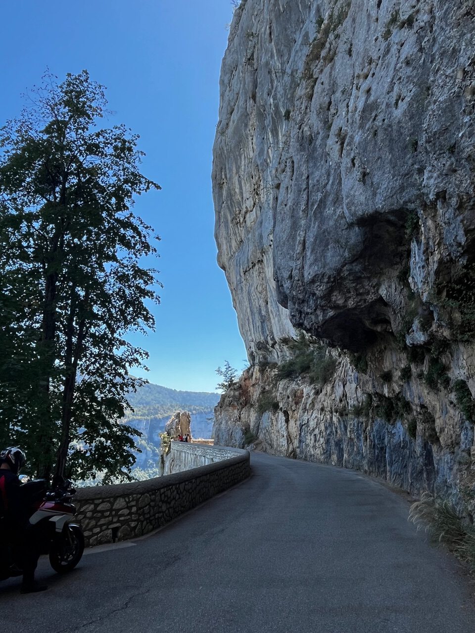 IMG_7696 Eine kurvenreiche Bergstraße schlängelt sich unter blauem Himmel an einer felsigen Klippe entlang; links sind ein Baum und ein Motorrad zu sehen.