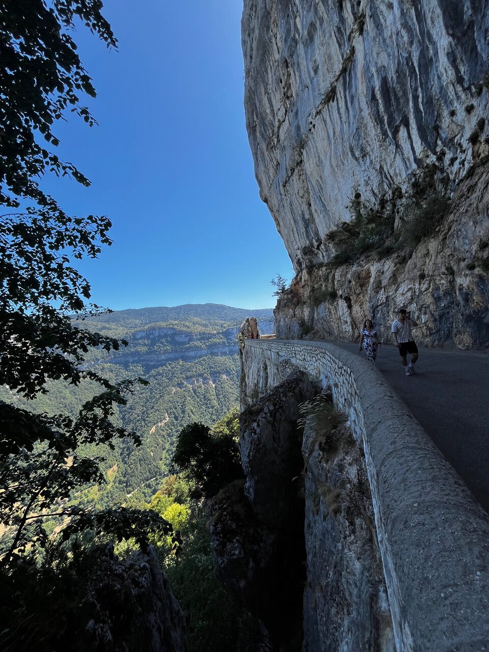 IMG_7697 Eine schmale Bergstraße schlängelt sich entlang einer steilen Felswand, Menschen stehen neben einer Steinmauer, darunter liegen üppig grüne Täler.