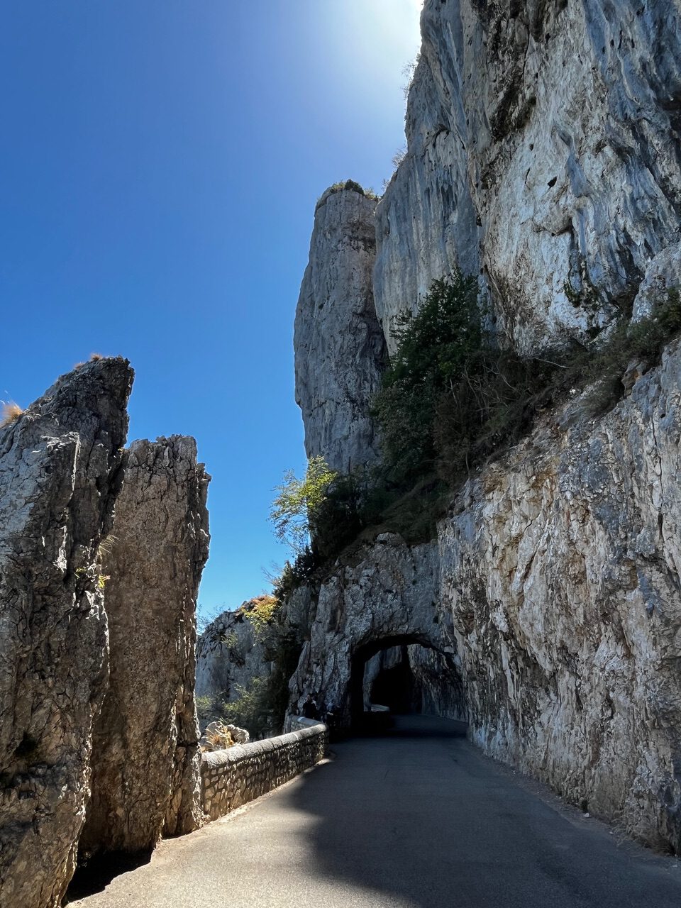 IMG_7699 Eine schmale Straße schlängelt sich durch einen Tunnel in den felsigen Bergen, Sonnenlicht und Schatten auf der Fahrbahn, darüber der blaue Himmel.