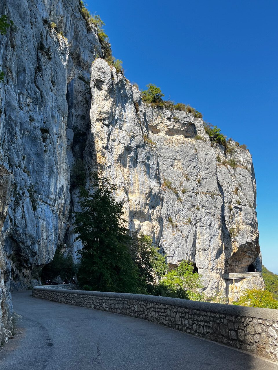IMG_7706 Eine kurvenreiche Straße schlängelt sich an einer hohen, felsigen Klippe entlang, die unter dem strahlend blauen Himmel mit grünen Flecken übersät ist.