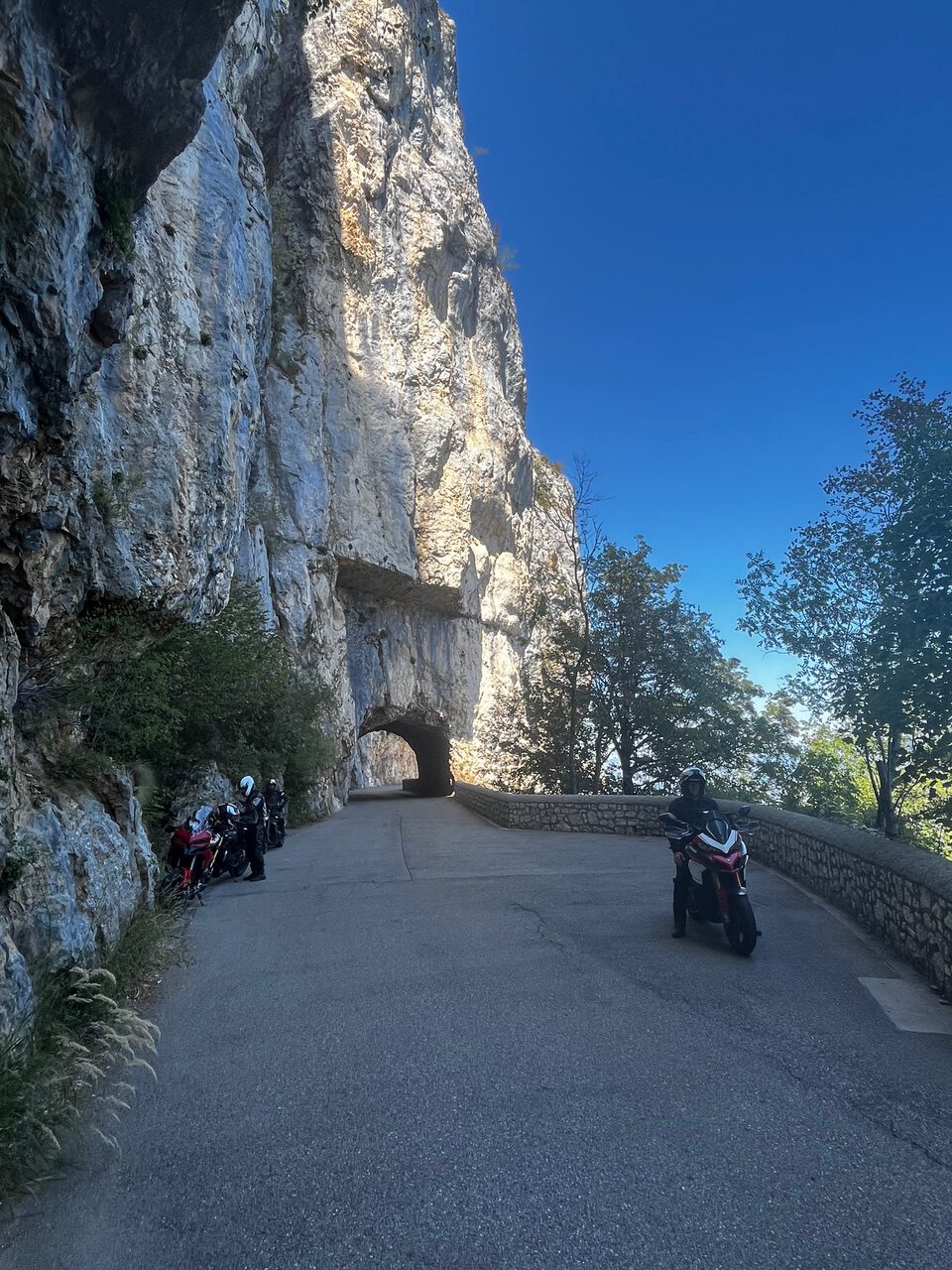 IMG_7707 Motorradfahrer halten auf einer schmalen Bergstraße neben einem felsigen Tunnel an; der Himmel ist klar, und am Straßenrand stehen Bäume.