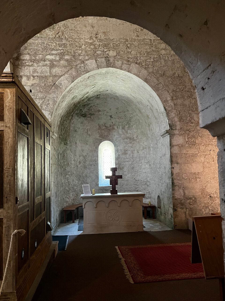 IMG_7718 Eine Kirche mit Steinmauern, Gewölbedecke, einem kleinen Altar und einem Kreuz, einem Bogenfenster und Holzbänken.
