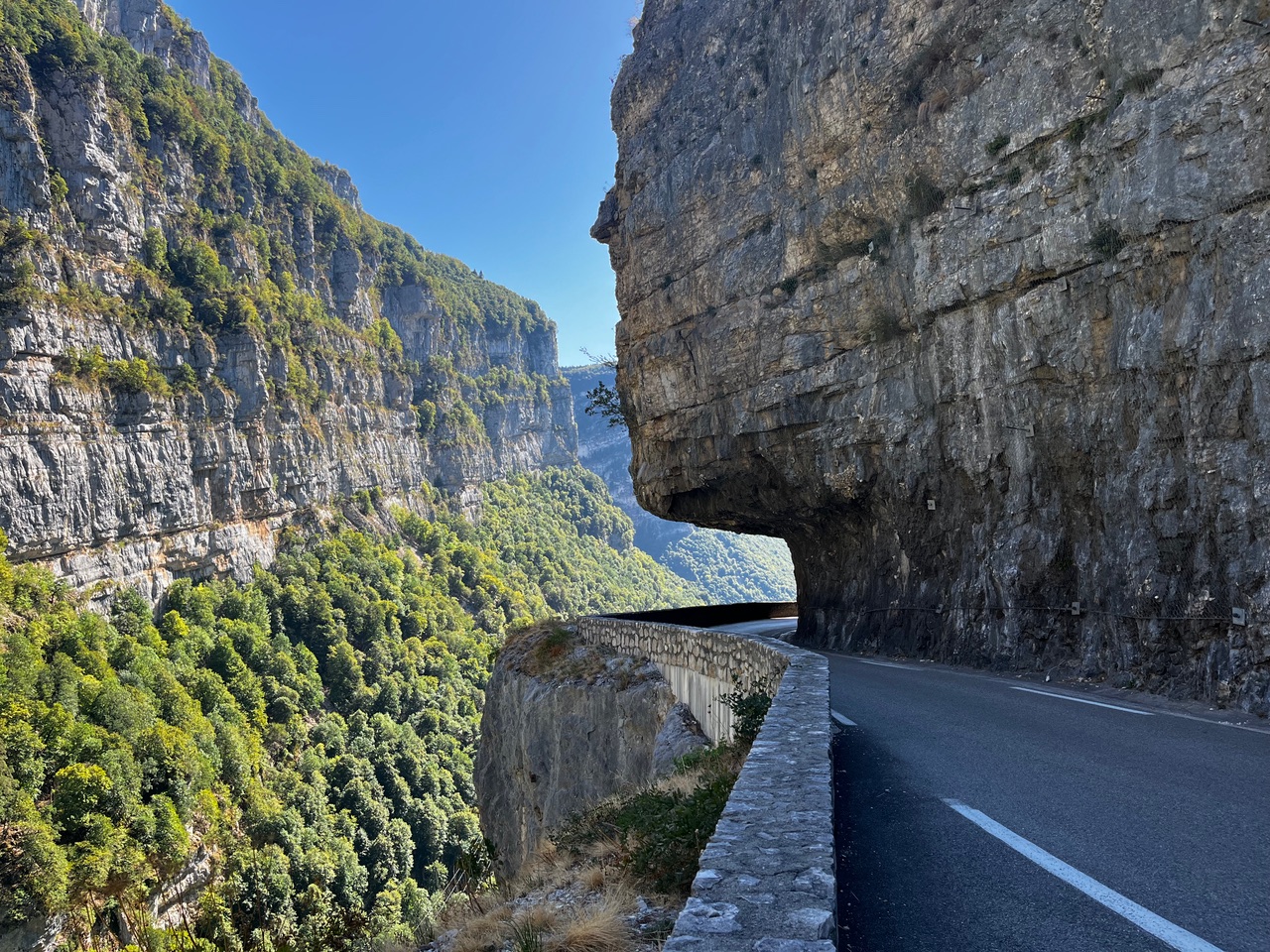 IMG_7729 Eine kurvenreiche Bergstraße führt unter einem gewaltigen Felsvorsprung hindurch, flankiert von bewaldeten Hängen und blauem Himmel.